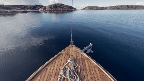 Du ser fören på en segelbåt i trä, du navigerar i ett kav lugnt skärgårdslandskap.